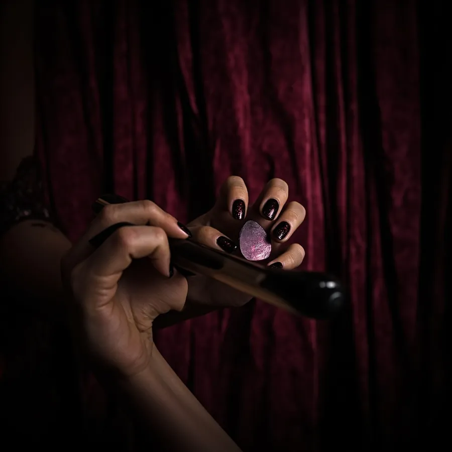 A hand with dark black cherry polished nails holding a small black brush and a pink heart shaped piece in front of a burgundy velvet curtain.