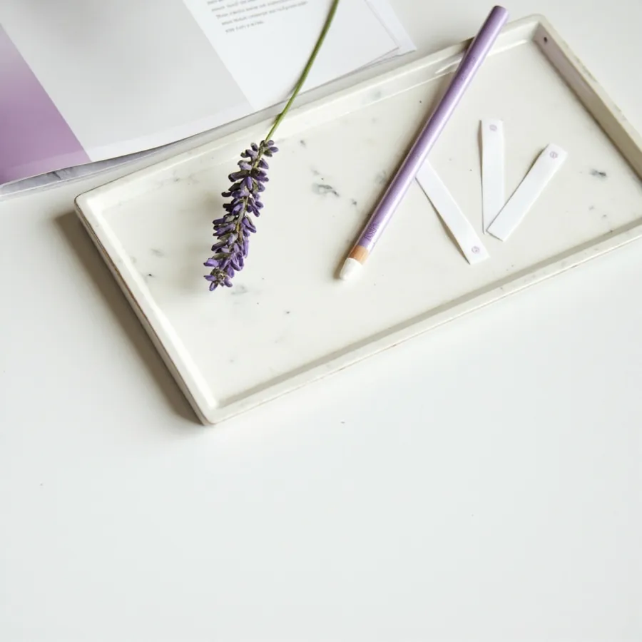 A lilac brow pencil, a sprig of lavender and two small white wax strips on a white marble tray.