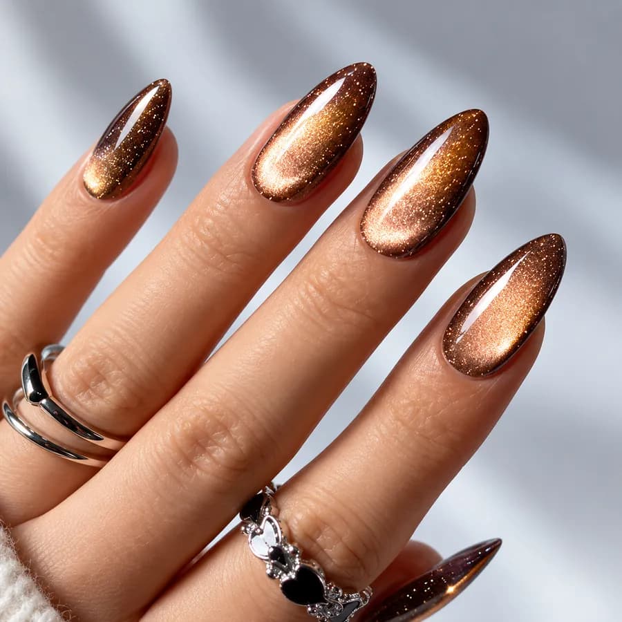 Long almond nails in glittering bronze cat eye gel, each nail with a warm band of copper light and fine gold sparkle, on a hand wearing silver rings.