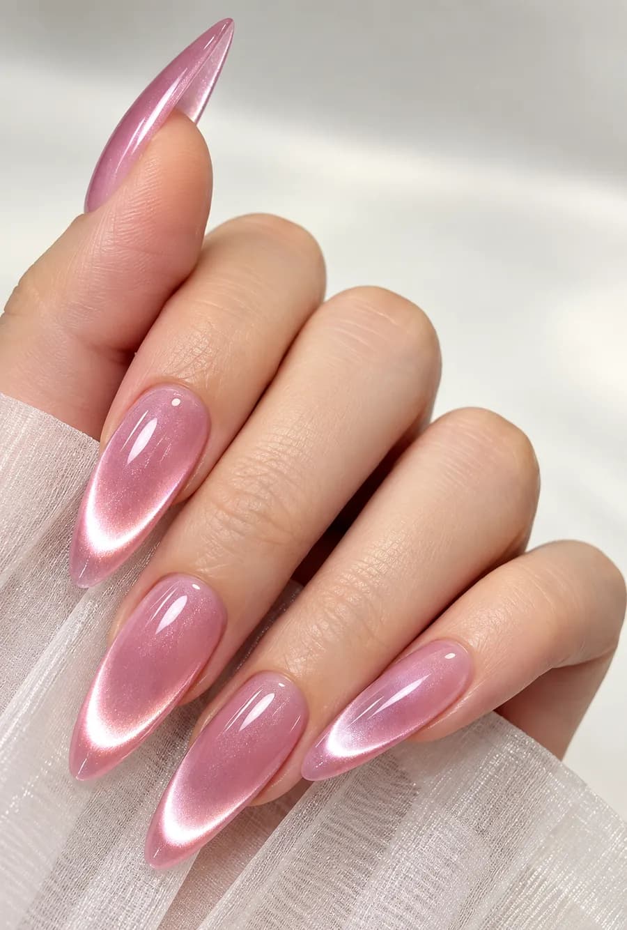 Long almond nails in pink aurora cat eye gel, each nail showing a soft white glowing arc of light, on a hand resting on sheer white fabric.